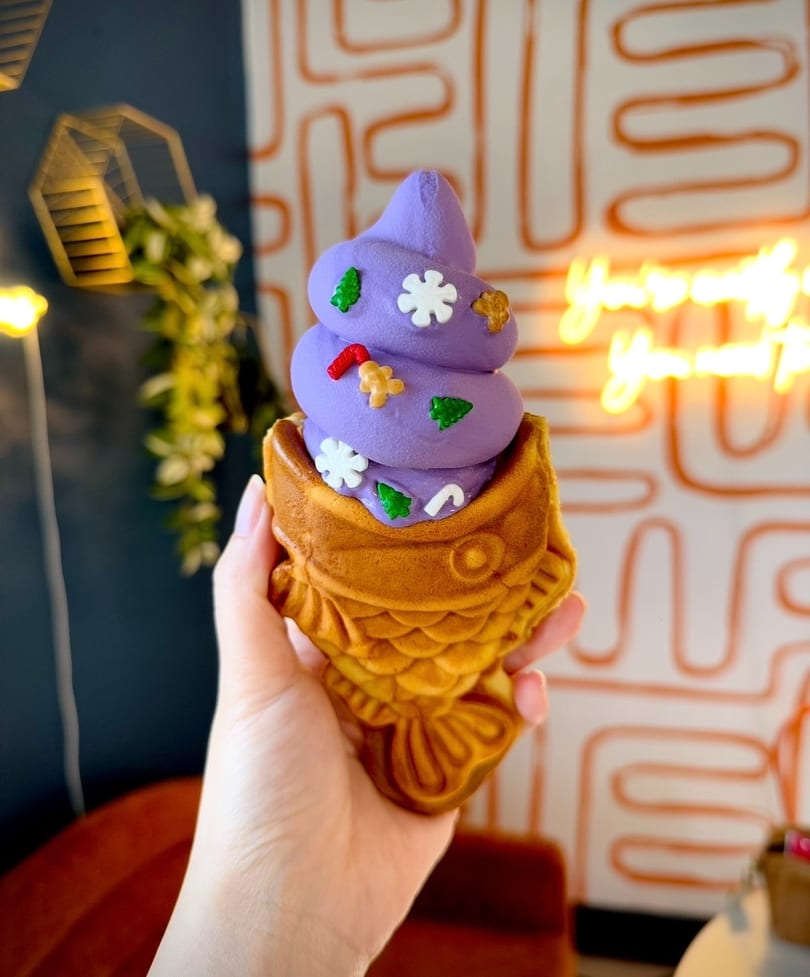 Hand holding purple soft-serve ice cream in a fish-shaped waffle cone with holiday sprinkles.