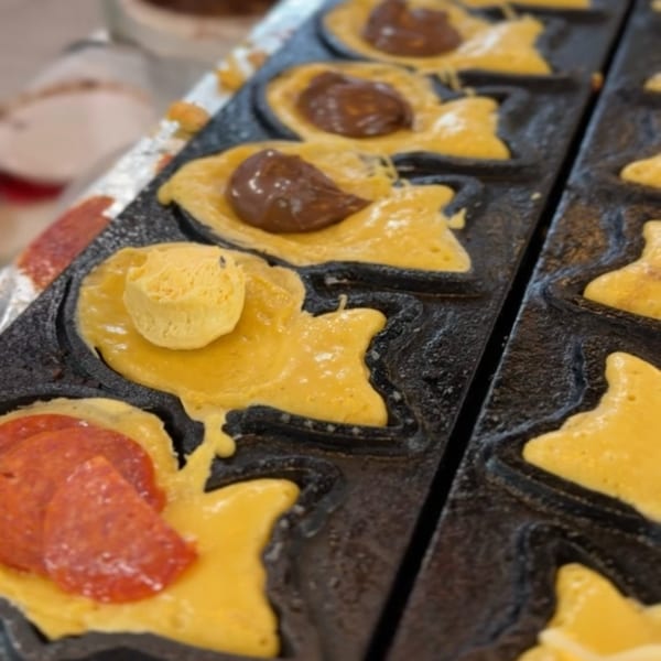 Fish-shaped taiyaki batter cooking in a mold with chocolate, pepperoni, and cheese fillings.