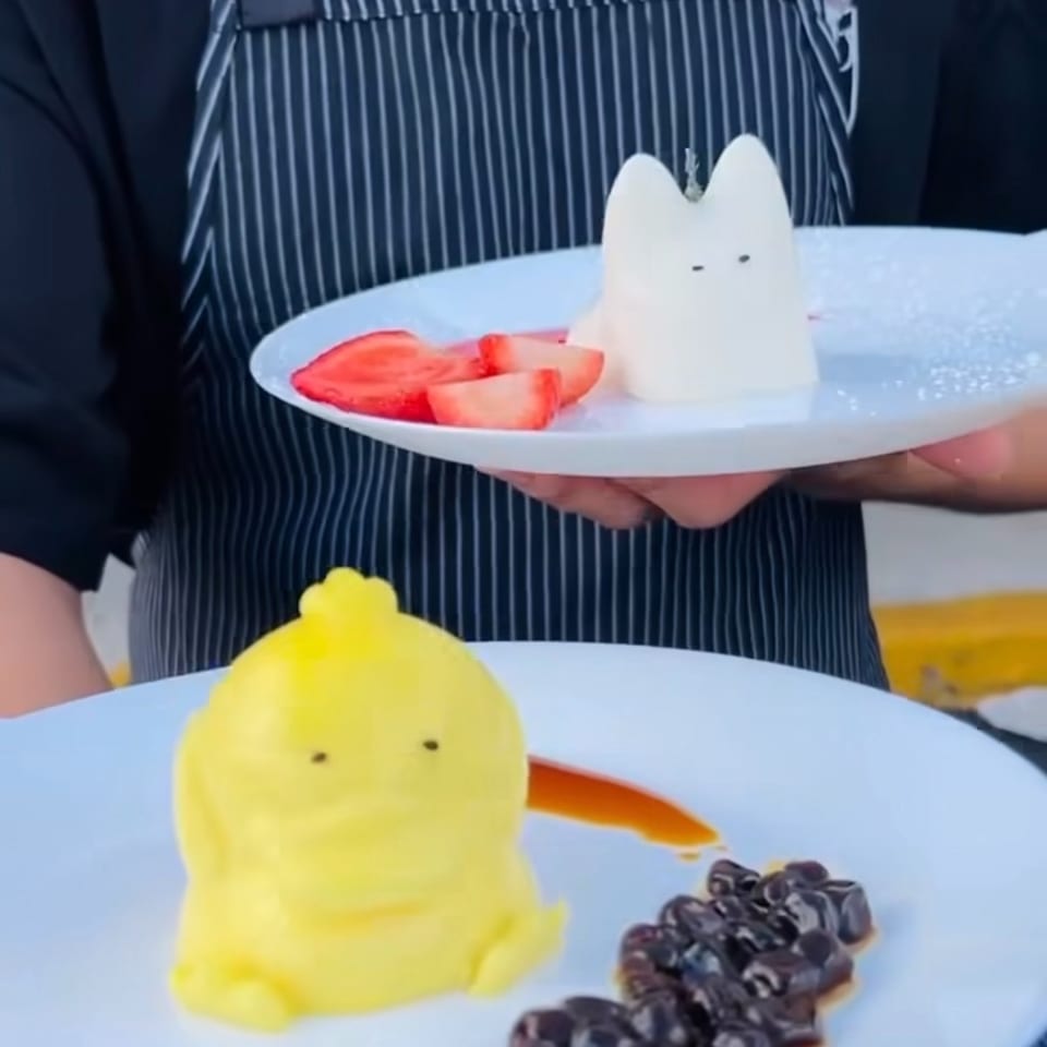 Chef holding two plates of whimsical desserts: a yellow character with boba and a white cat with strawberries.