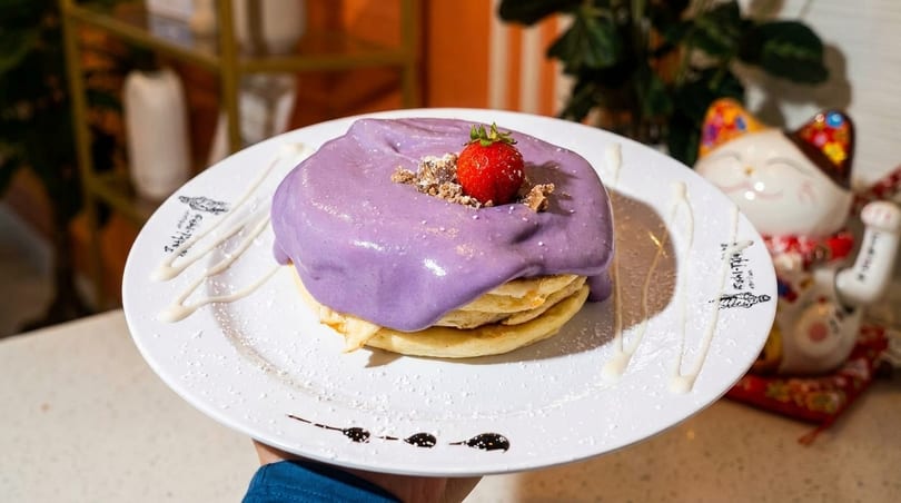 Fluffy pancakes topped with vibrant purple ube cream, a fresh strawberry, and cookie crumbles.