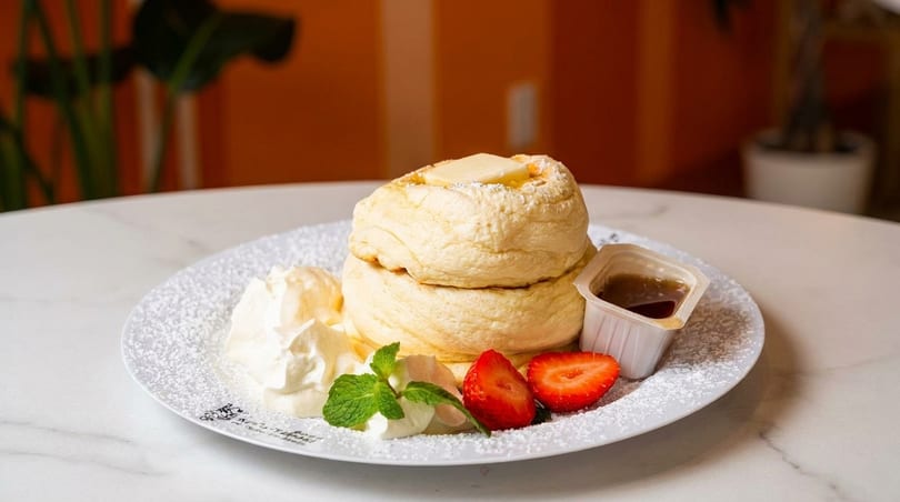 Fluffy soufflé pancakes topped with butter, whipped cream, strawberries, and syrup on a white plate.