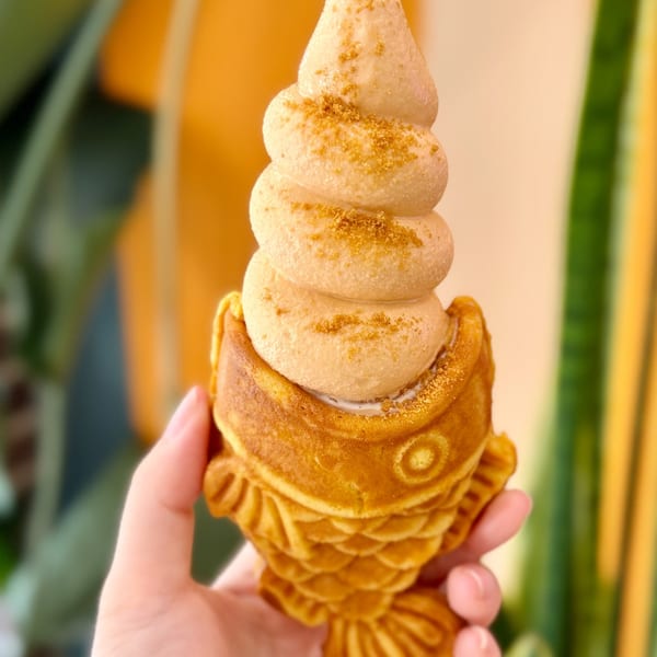 Golden fish-shaped Taiyaki waffle cone filled with swirled soft-serve ice cream, held by a hand.