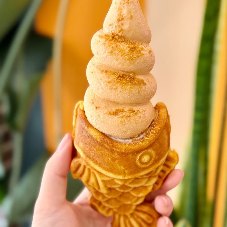 Golden fish-shaped Taiyaki waffle cone filled with swirled soft-serve ice cream, held by a hand.