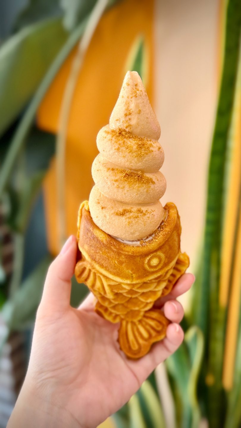 Golden fish-shaped Taiyaki waffle cone filled with swirled soft-serve ice cream, held by a hand.