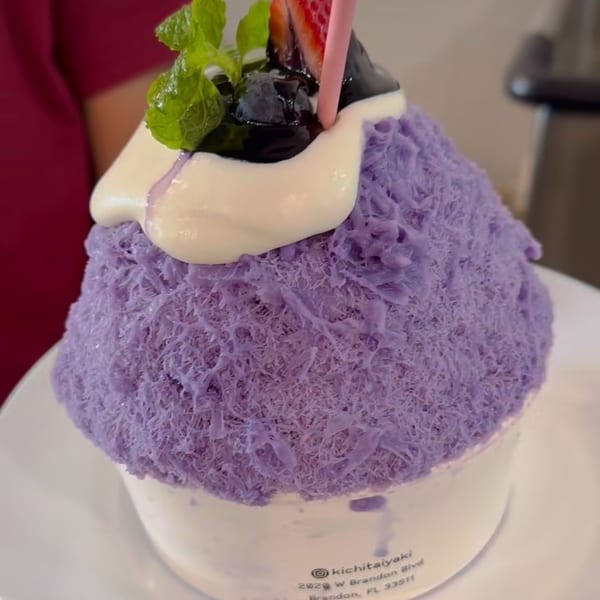 Purple shaved ice dessert topped with cream, blueberries, strawberry, mint, and a pink straw.