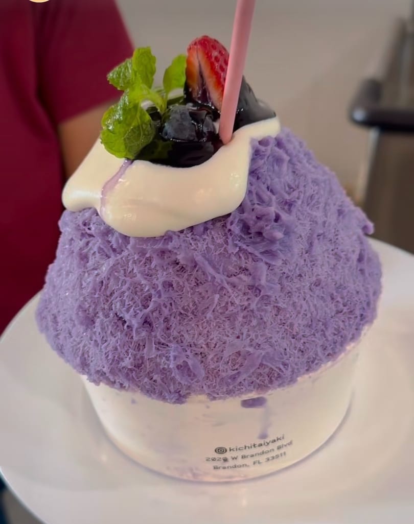 Purple shaved ice dessert topped with cream, blueberries, strawberry, mint, and a pink straw.