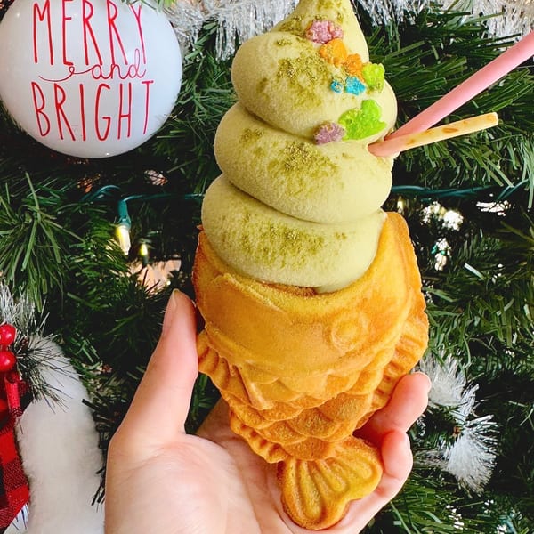 Hand holds matcha soft serve in a fish-shaped taiyaki cone with sprinkles and pocky sticks, against a Christmas tree.