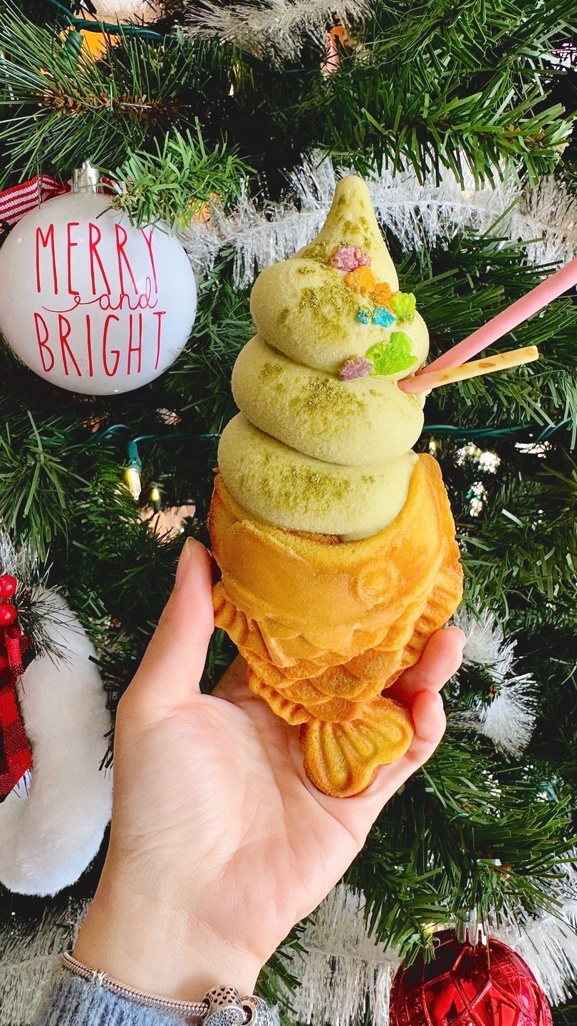 Hand holds matcha soft serve in a fish-shaped taiyaki cone with sprinkles and pocky sticks, against a Christmas tree.
