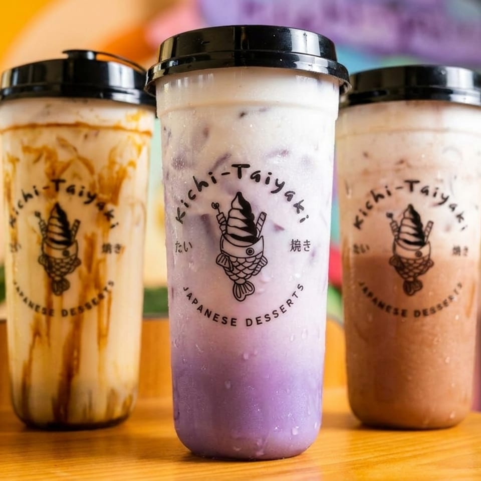 Three Kichi-Taiyaki bubble tea drinks, including taro, on a wooden counter with a colorful background.