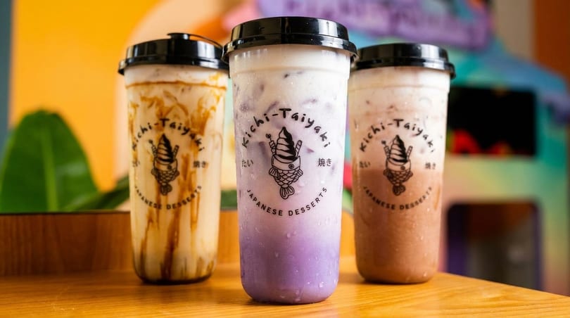 Three Kichi-Taiyaki bubble tea drinks, including taro, on a wooden counter with a colorful background.